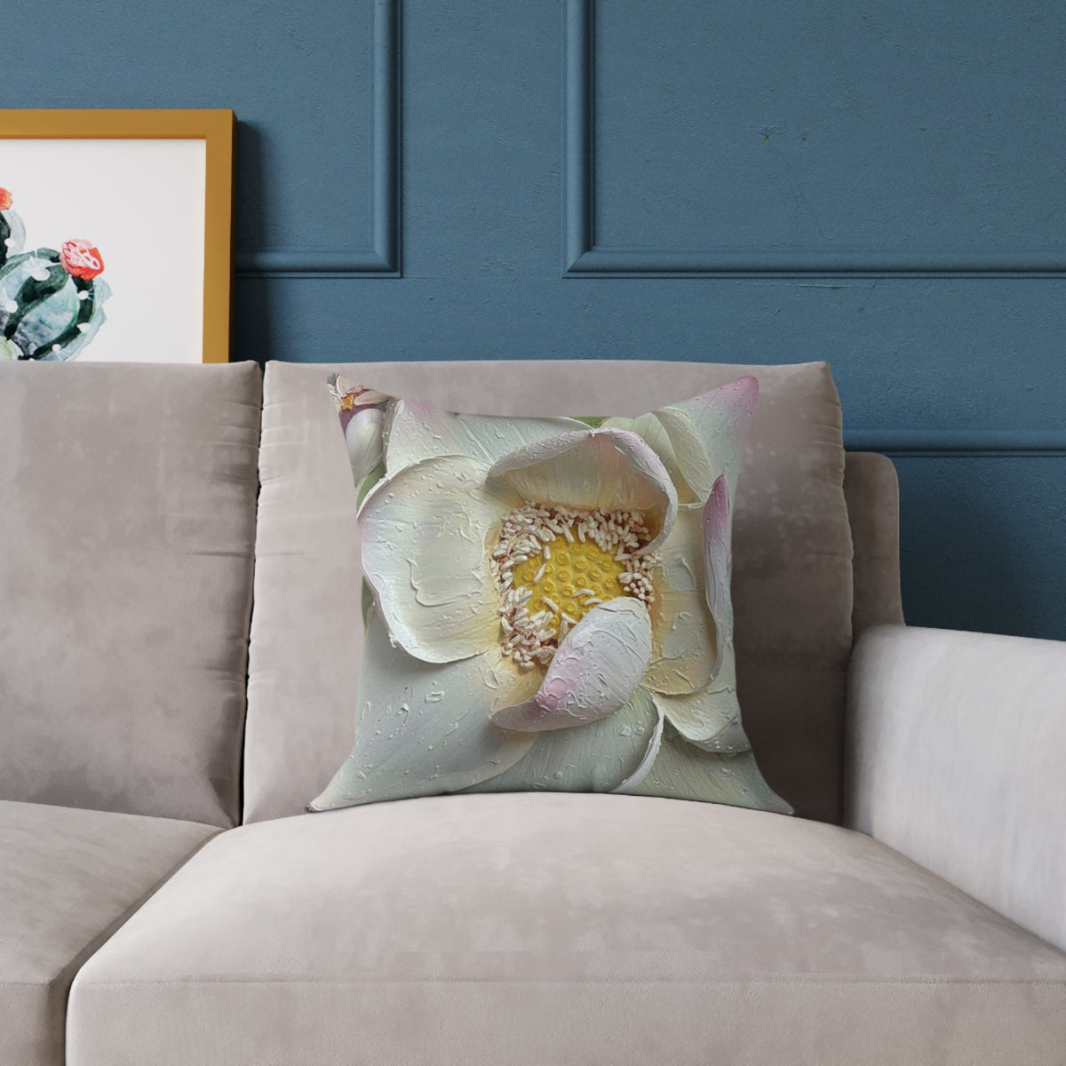 A square throw pillow featuring a close-up print of a pale pink and white lotus flower with a yellow center, resting on a beige velvet sofa against a dark blue paneled wall.
