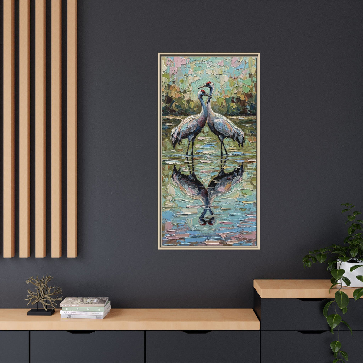A framed vertical painting of two brolgas standing in reflective water, displayed on a dark charcoal wall above a light wood console with books and a plant.
