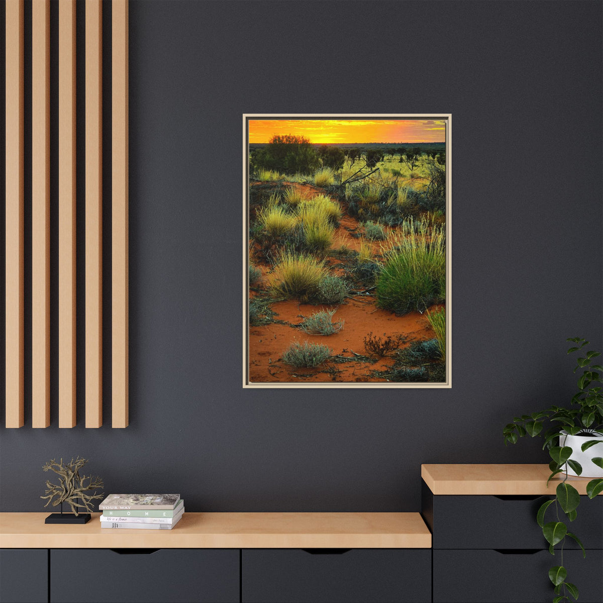 A vertical framed art print depicting a desert landscape at sunset hangs on a dark charcoal wall above a light wood console table with decorative items and a plant.