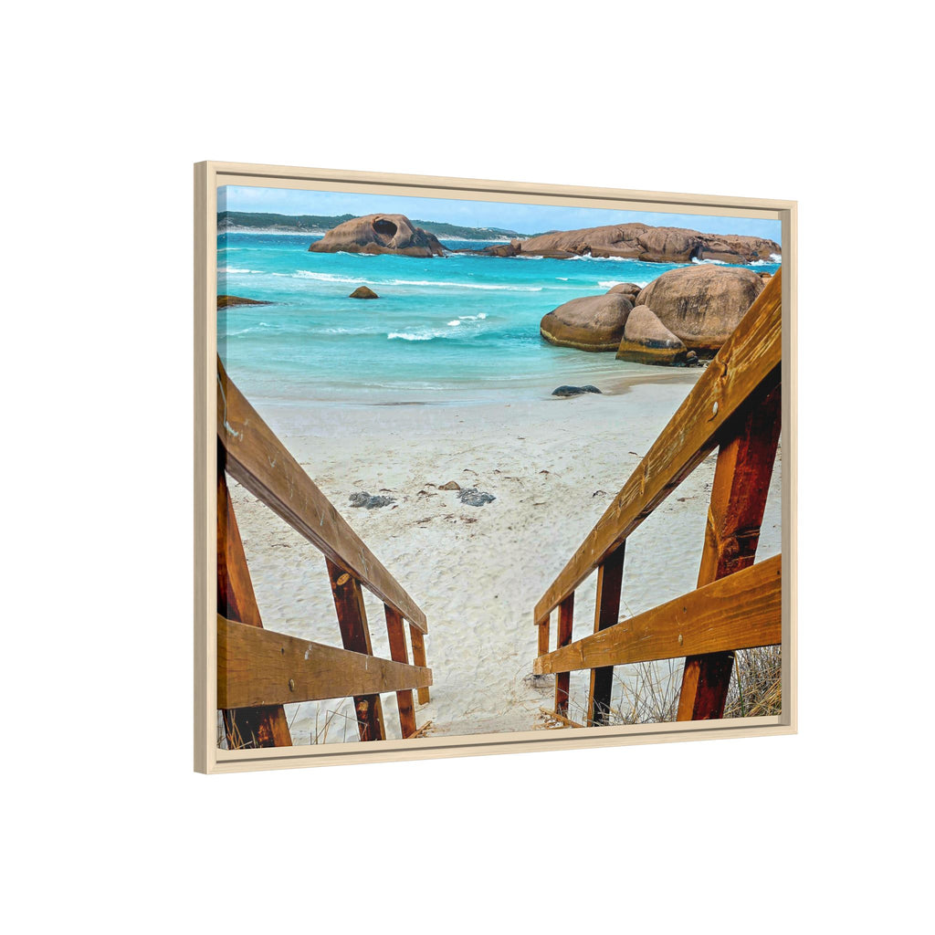 Framed square wall art showing a coastal beach scene with turquoise water, large rocks, pale sand, and wooden stairs leading into the frame, displayed against a light gray background.