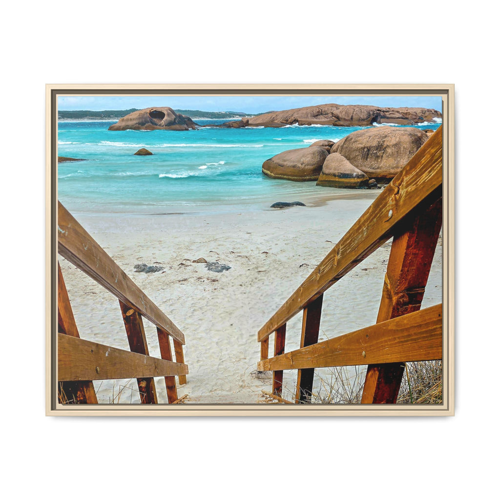 A framed photographic print of a coastal beach scene, viewed from above through wooden stairs toward a sandy shore, turquoise ocean water, and large rock formations. The print is presented in a light wood frame against a plain white background.