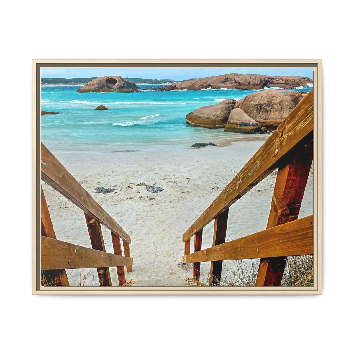 A framed photographic print of a coastal beach scene, viewed from above through wooden stairs toward a sandy shore, turquoise ocean water, and large rock formations. The print is presented in a light wood frame against a plain white background.