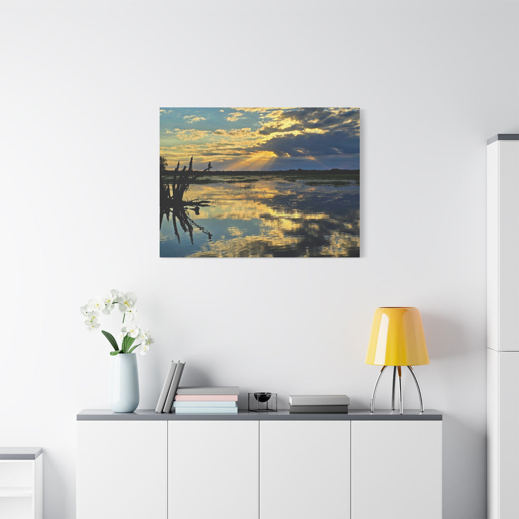 A square wall art print showing a sunset over calm water with reflections and dark tree branches on the left, displayed above a white cabinet in a modern room with a yellow lamp and white flowers.