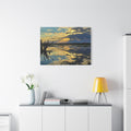 A square wall art print showing a sunset over calm water with reflections and dark tree branches on the left, displayed above a white cabinet in a modern room with a yellow lamp and white flowers.