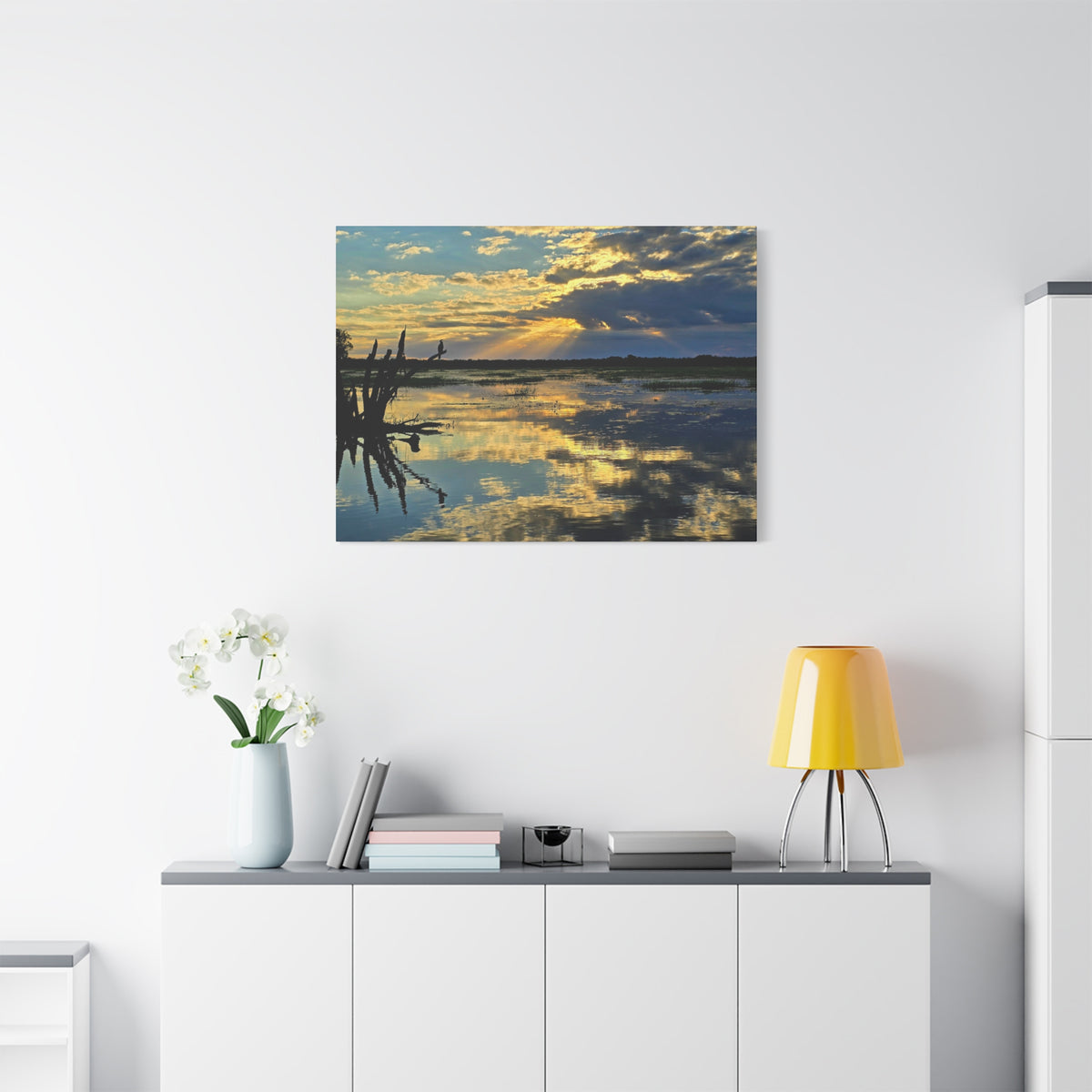 A square wall art print showing a sunset over calm water with reflections and dark tree branches on the left, displayed above a white cabinet in a modern room with a yellow lamp and white flowers.