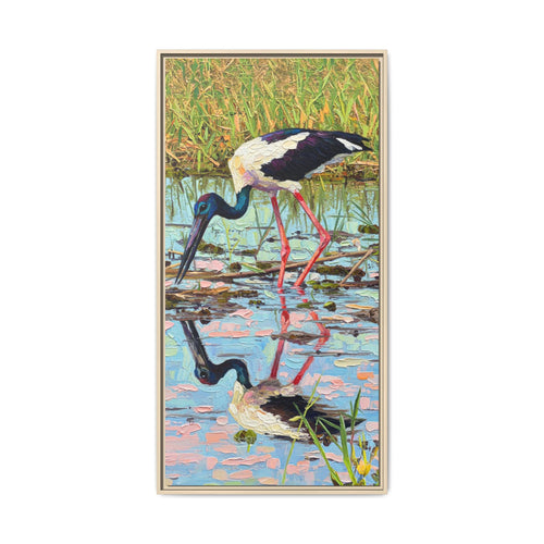 A framed vertical art print depicting a black-necked stork standing in shallow water with its reflection visible, set against a plain white background.