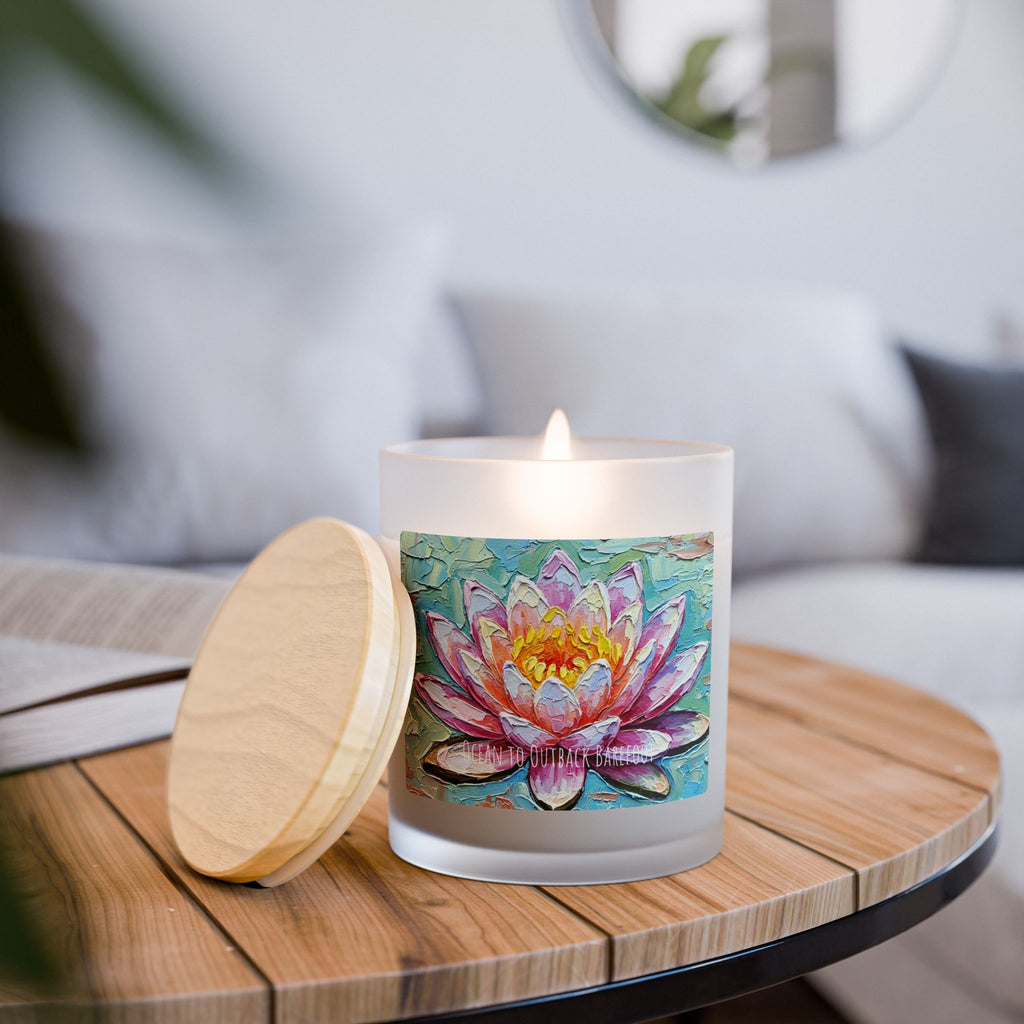 A frosted glass candle with a lit flame sits on a round wooden table, next to its light wood lid. The candle features a colorful water lily illustration on the front.