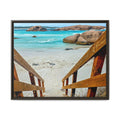 A framed photographic print of a coastal beach scene, viewed from above through wooden steps onto pale sand, turquoise water, and large rock formations. The artwork is presented in a dark frame against a plain white background.