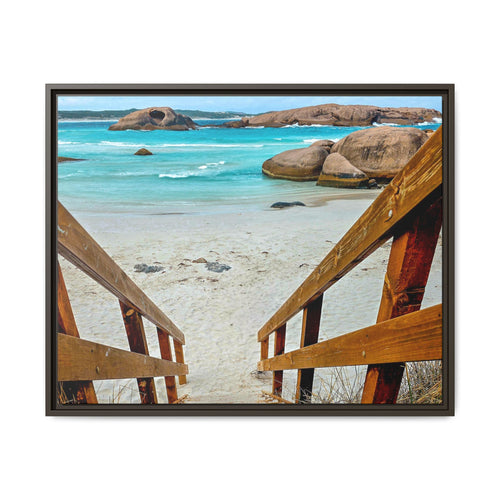 A framed photographic print of a coastal beach scene, viewed from above through wooden steps onto pale sand, turquoise water, and large rock formations. The artwork is presented in a dark frame against a plain white background.
