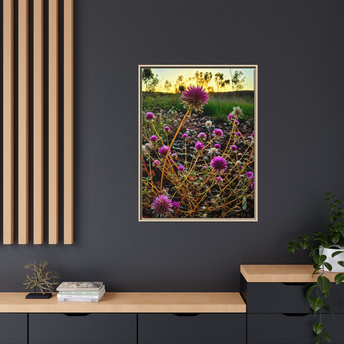 A vertical framed art print depicting purple bachelor button flowers in a field at sunset, displayed on a dark gray wall above a wooden console table in a modern room setting.