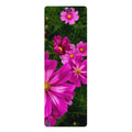 A vertical yoga mat featuring a vibrant print of pink cosmos flowers with yellow centers against a dark green leafy background. The word 'Yoga' is printed in black at the top, and a small circular blue logo is at the bottom left.