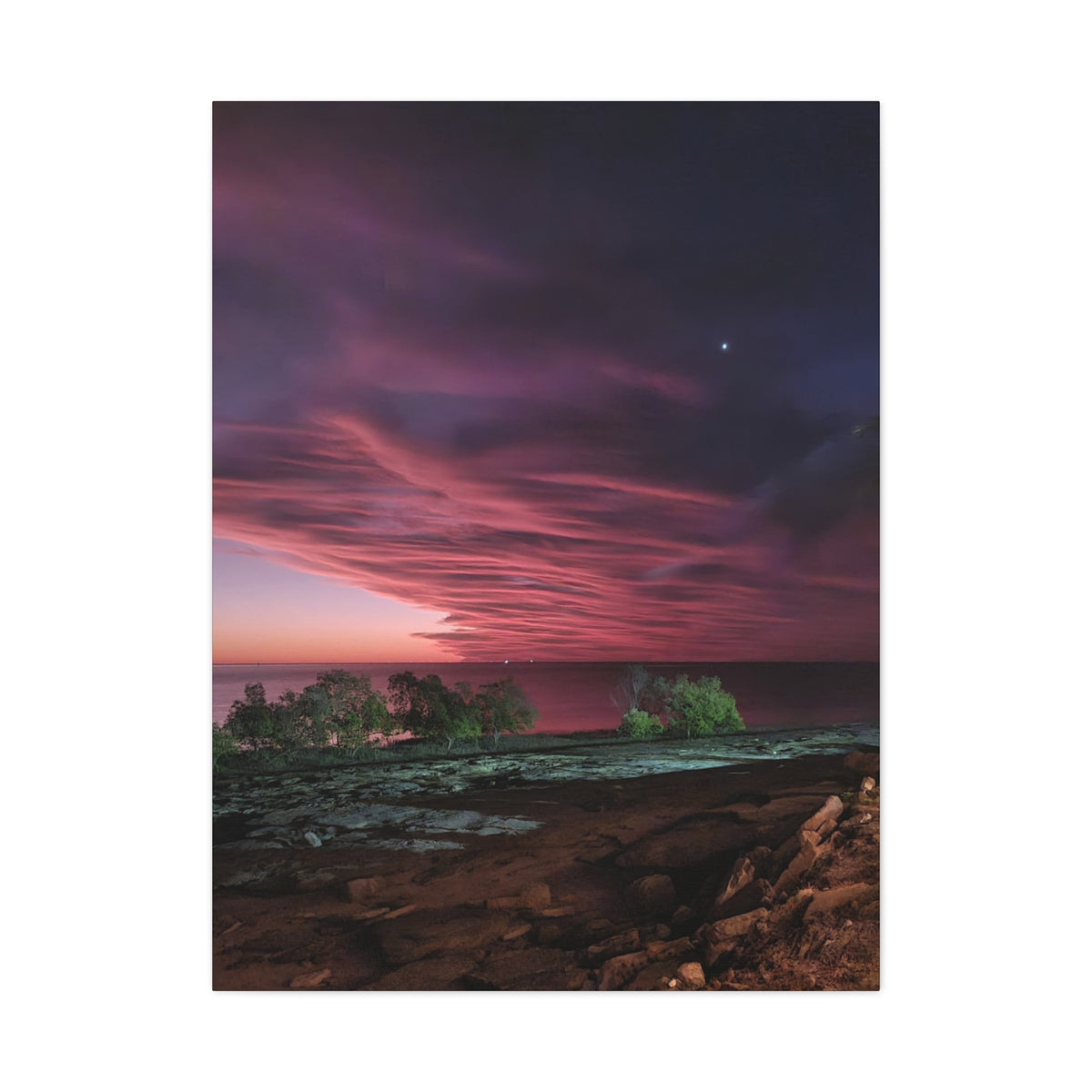 A vertical photographic art print depicting a dramatic twilight seascape with a dark purple and pink sky, glowing clouds, a horizon line, rocky shoreline, and small green trees in the water.