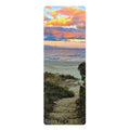 A tall rectangular yoga mat featuring a scenic beach landscape print with a sunset sky, ocean, dunes, and a stone path. The word 'Yoga' is printed near the bottom, and a small circular logo appears in the lower left corner.