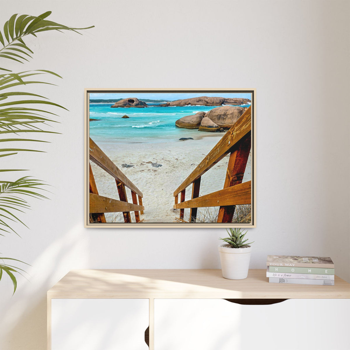 A framed square wall art print showing a tropical beach scene with turquoise water, white sand, large rocks, and wooden stair railings leading down to the shore, displayed on a white wall above a light wood console table with a small plant and books.