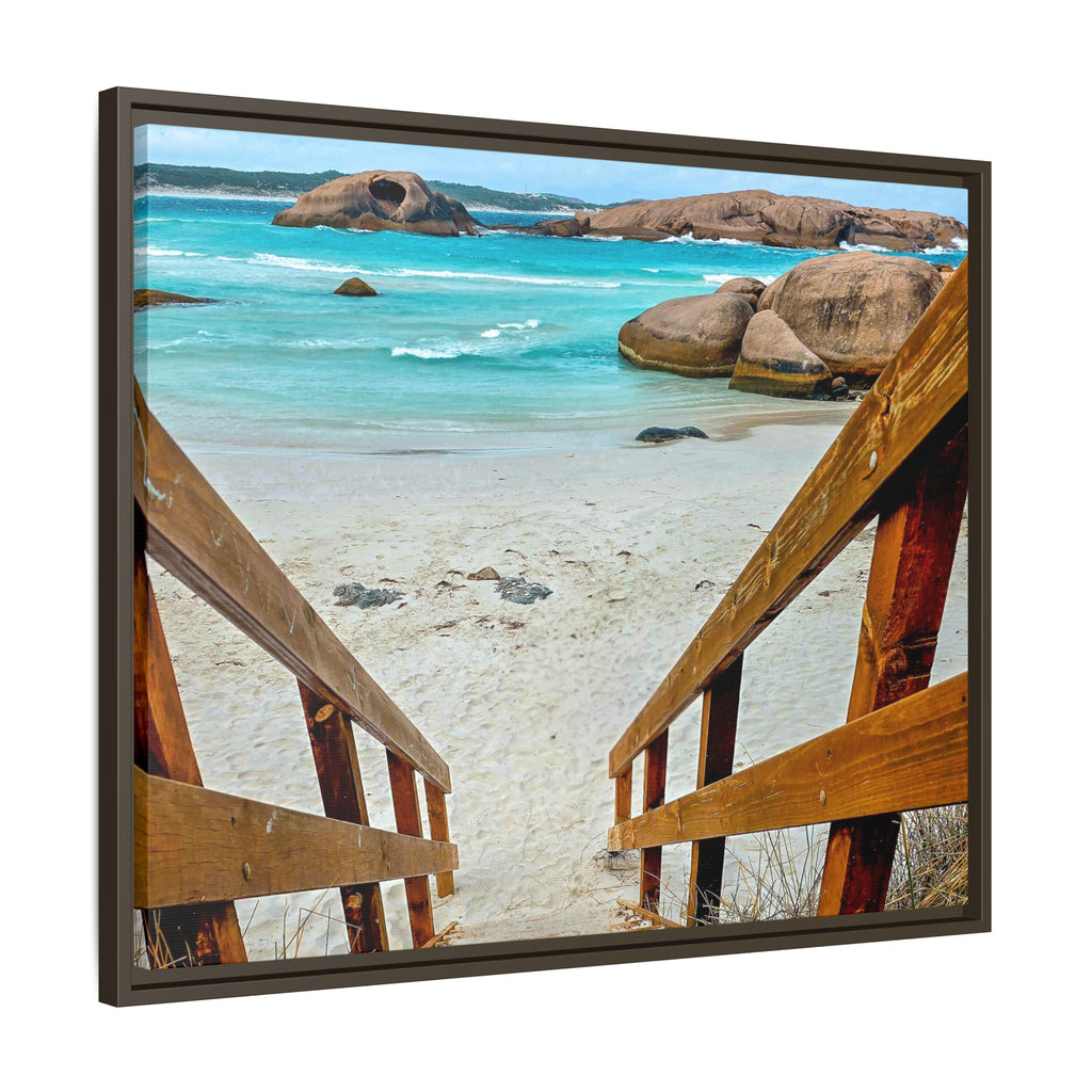 Framed square wall art showing a beach scene viewed from wooden stairs, with turquoise water, sandy shore, large rock formations, and a dark wood frame on a white background.