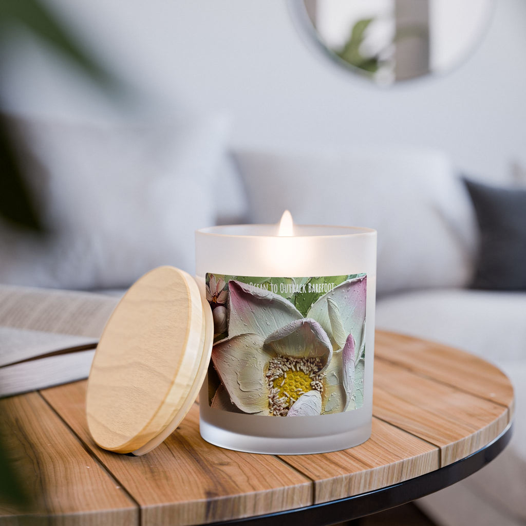 A lit frosted glass candle with a colorful lotus flower label sits on a round wooden table next to its light wood lid in a softly blurred living room setting.