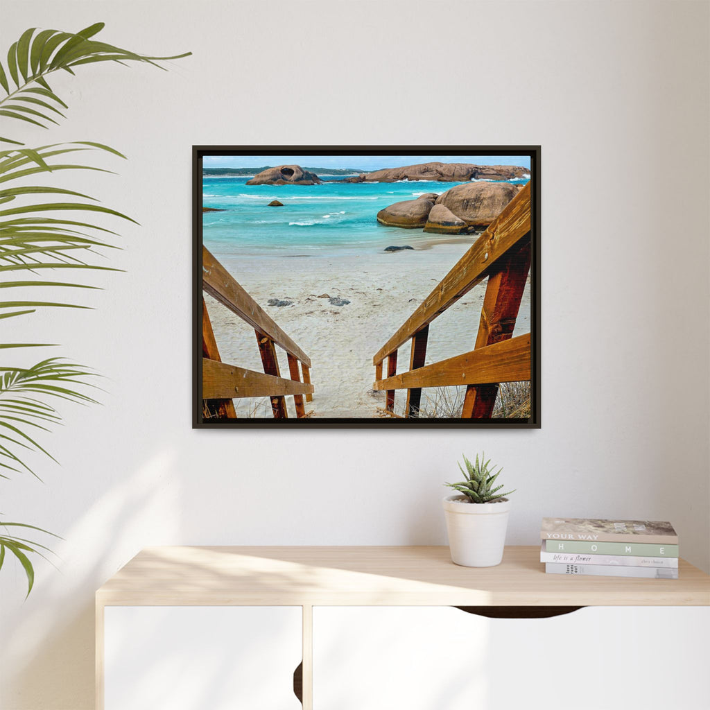 A framed square wall art print showing a beach scene viewed from wooden stairs, displayed on a white wall above a light wood console table with a small plant and books.