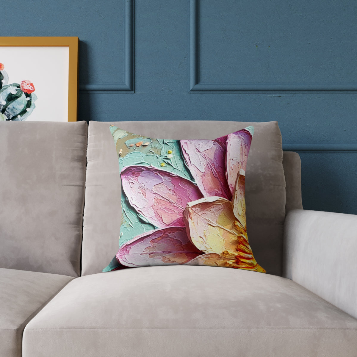 A square throw pillow featuring a colorful, textured oil-painted lotus flower design in shades of pink, purple, and yellow, styled on a beige sofa against a dark blue paneled wall.