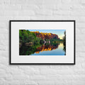 A framed landscape art print of a scenic river reflecting orange and yellow cliffs, hung on a white brick wall.