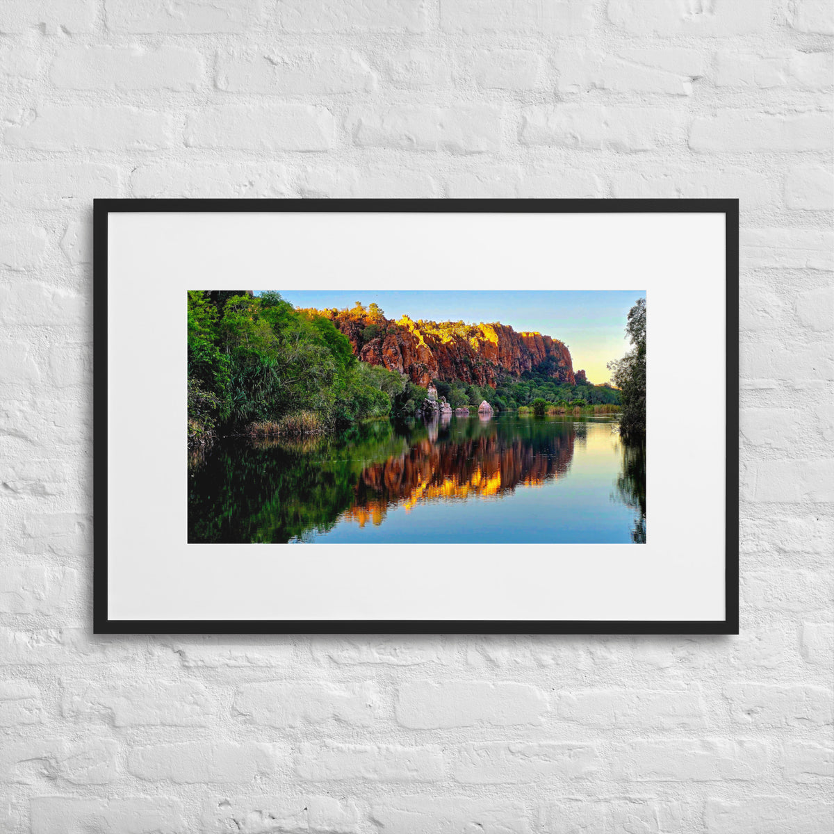 A framed landscape art print of a scenic river reflecting orange and yellow cliffs, hung on a white brick wall.