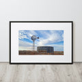 A framed landscape art print featuring a windmill and a water tank under a blue sky, displayed on a light gray wall above a wood-look floor.