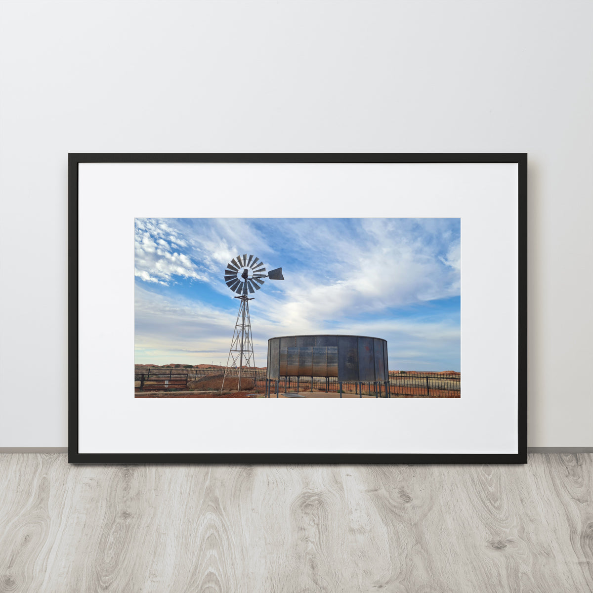 A framed landscape art print featuring a windmill and a water tank under a blue sky, displayed on a light gray wall above a wood-look floor.