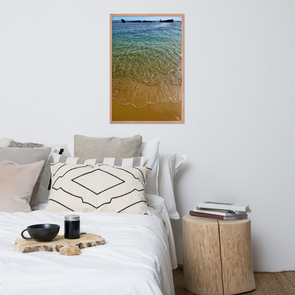 A framed vertical art print depicting a beach scene with turquoise water and golden sand, styled on a white wall above a bed in a modern, minimalist bedroom.