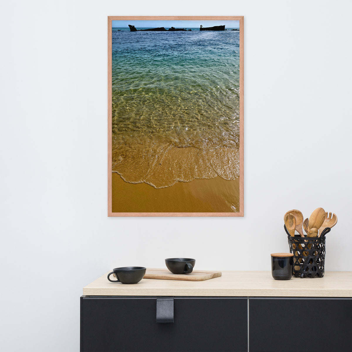 A vertical framed art print depicting a beach shoreline with turquoise water, golden sand, and small boats on the horizon, displayed on a white wall above a kitchen counter with black cups and utensils.