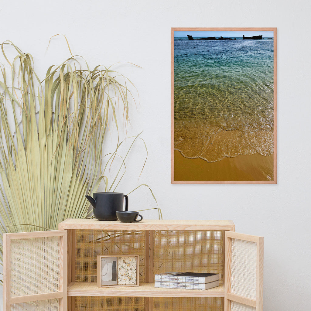 A framed vertical art print depicting turquoise ocean waves washing onto a sandy beach, styled on a white wall above a light wood cabinet with a potted plant and decorative items.