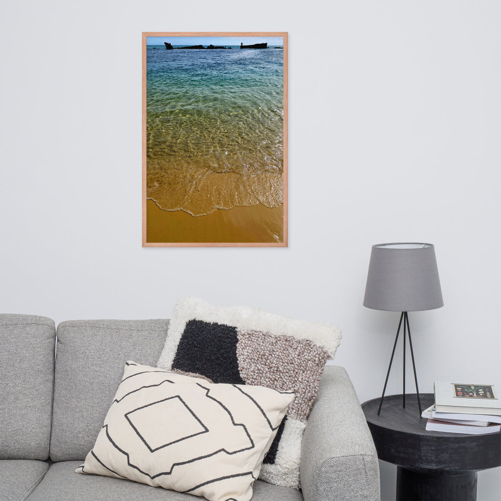 A vertical framed art print depicting a beach scene with turquoise water, golden sand, and small boats on the horizon, styled on a white wall above a gray sofa with decorative pillows and a side table with a lamp.