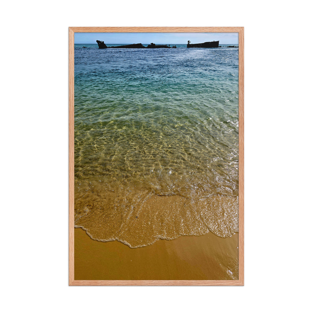 A framed photographic print of a beach scene with clear turquoise water, golden sand, and a distant shipwreck, set in a light natural wood frame against a white background.