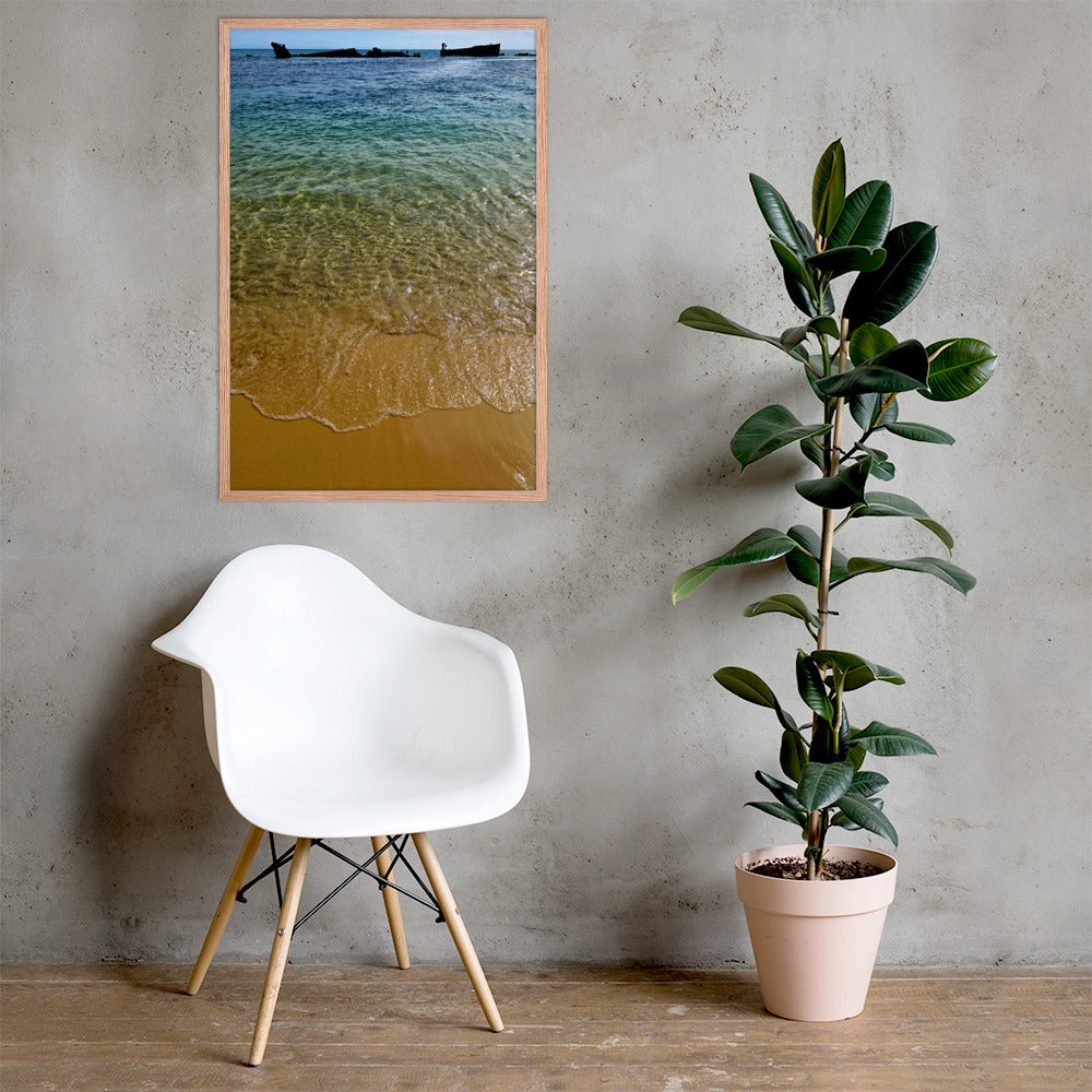 A framed vertical art print of a beach scene with turquoise water and golden sand hangs on a textured gray wall above a white molded chair and next to a potted rubber plant.