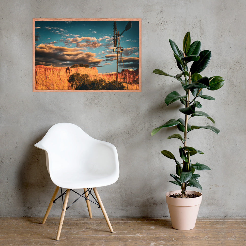 A framed landscape art print depicting a desert canyon at sunset with a windmill, displayed on a gray concrete wall in a modern room setting with a white chair and a potted rubber plant.