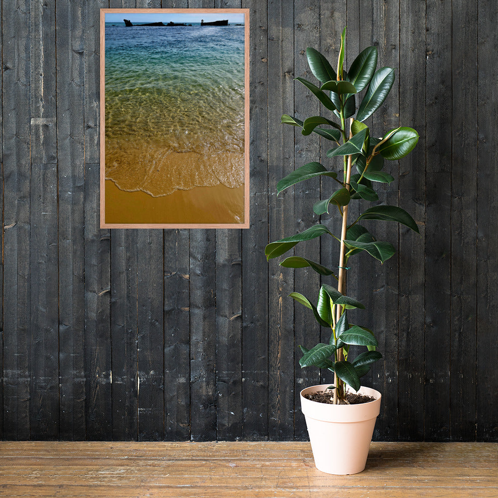 A framed vertical beach print showing turquoise water and golden sand, displayed on a dark wood plank wall next to a potted rubber plant.