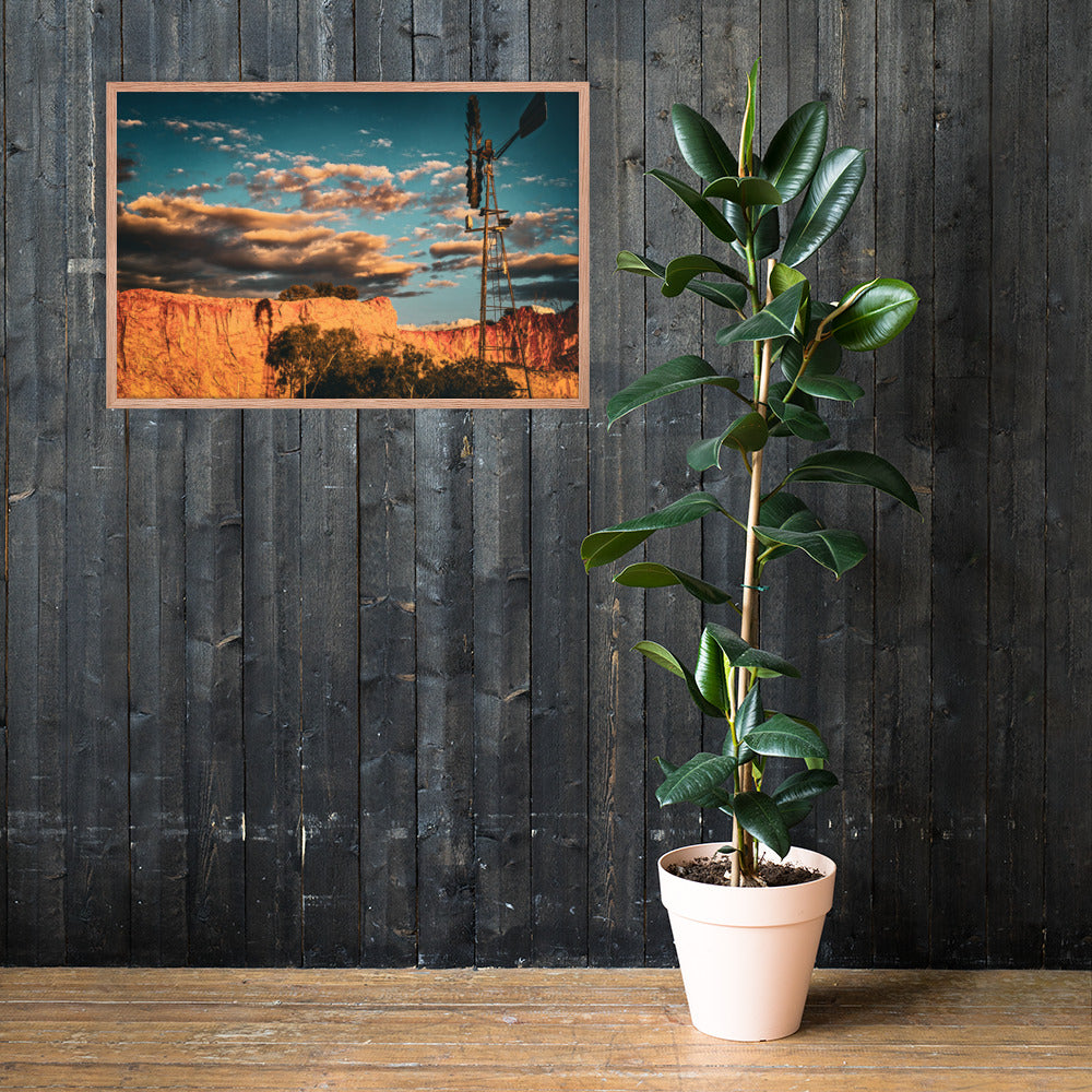 A framed landscape art print depicting a desert canyon at sunset with a windmill, displayed on a dark wood-paneled wall next to a potted rubber plant.