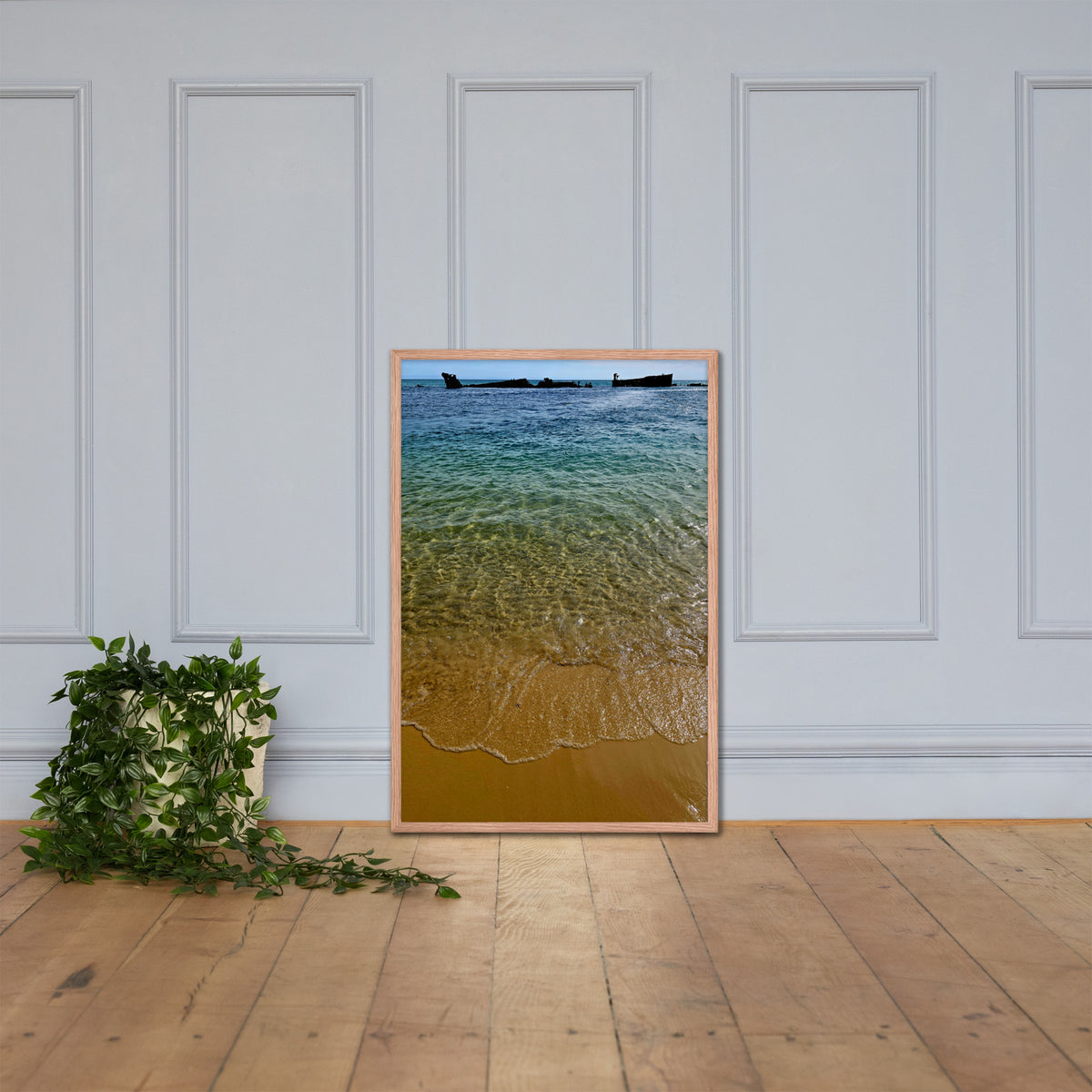 A vertical framed art print depicting a beach scene with clear turquoise water, golden sand, and small boats on the horizon, leaning against a white paneled wall on a light wood floor next to a green potted plant.