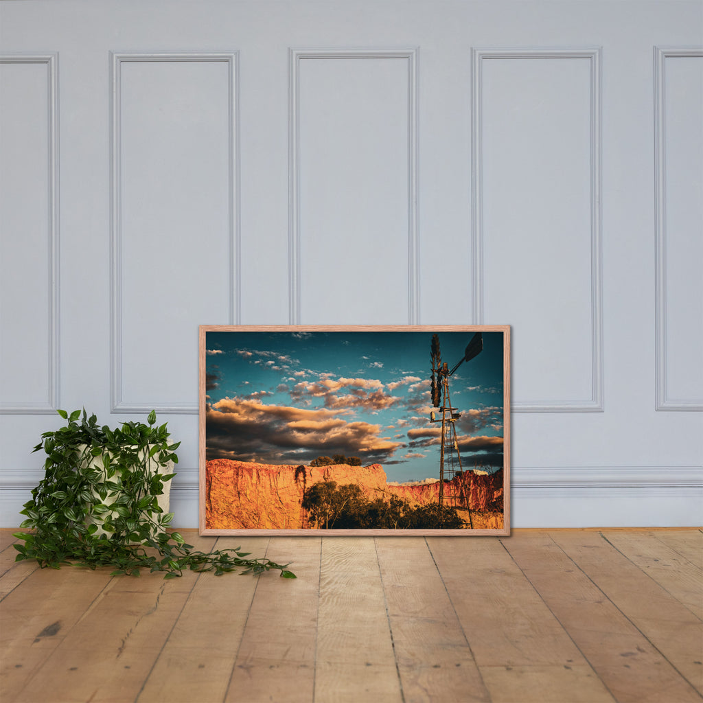 A framed landscape art print depicting a canyon at sunset with a windmill, leaning against a white paneled wall on a light wood floor next to a green potted plant.