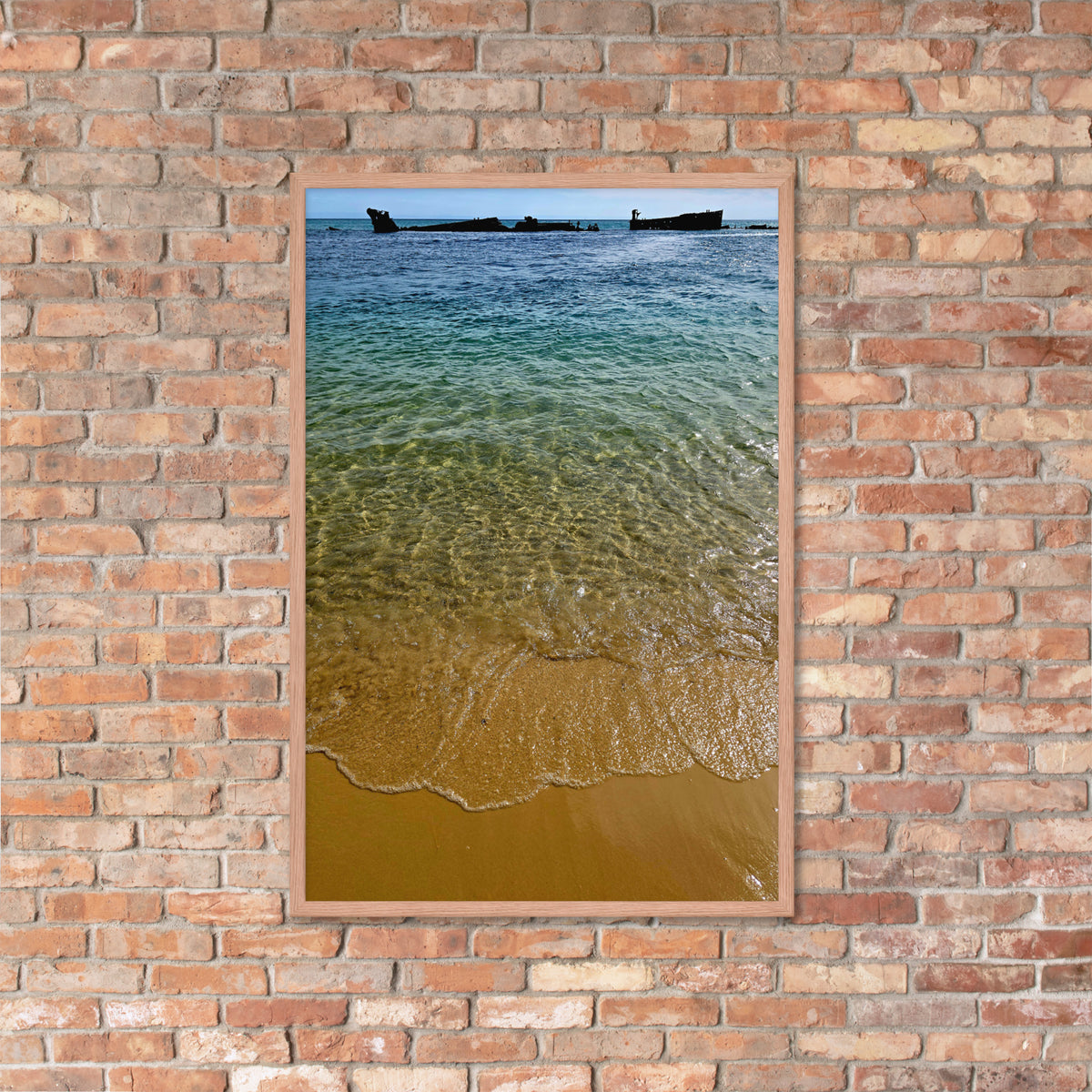 A vertical framed art print depicting a beach scene with clear turquoise water, golden sand, and small boats on the horizon, mounted on a rustic red-brown brick wall.