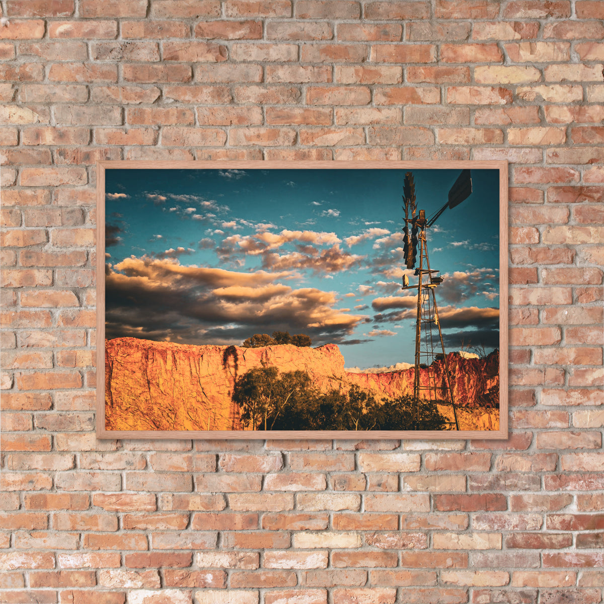 A framed landscape art print depicting a canyon scene with a windmill under a blue sky, mounted on a rustic brick wall.